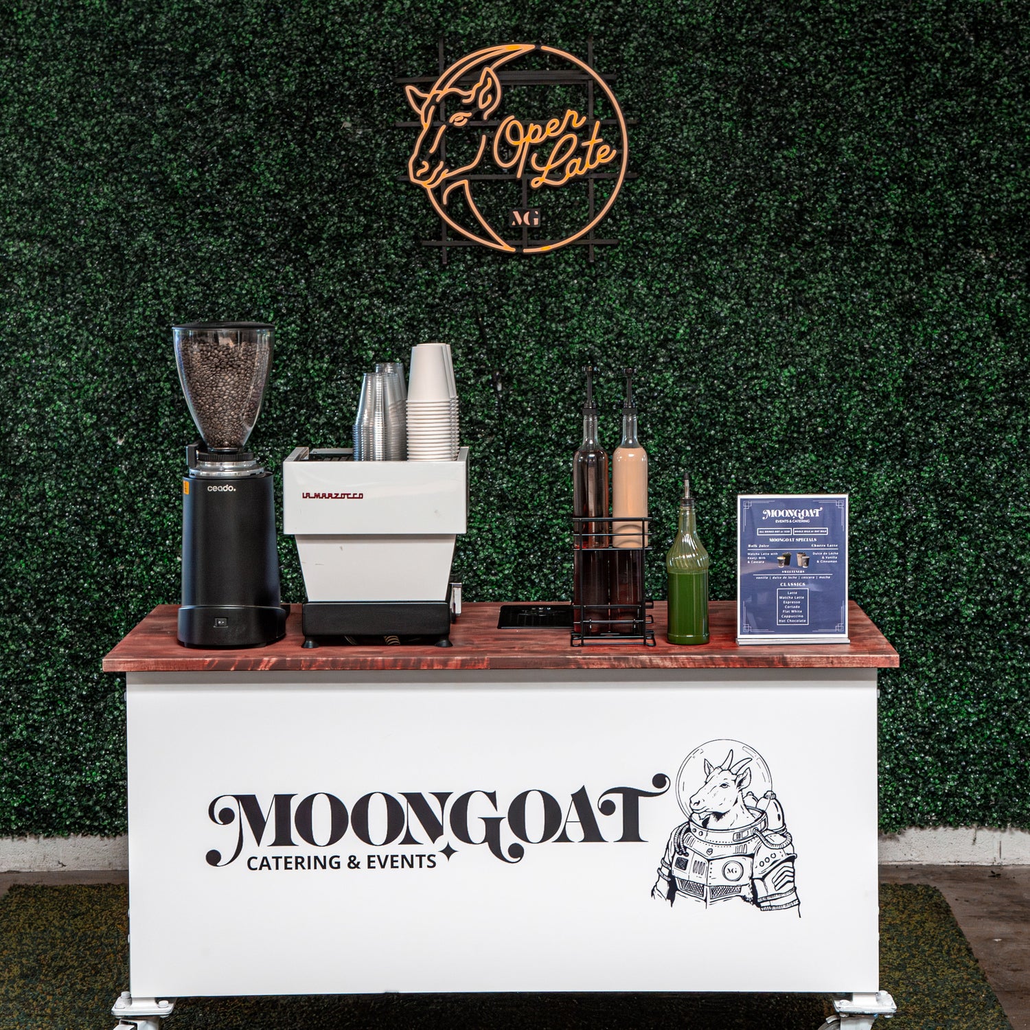 Catering cart with coffee equipment and branding against a green wall with 'Open Late' sign.